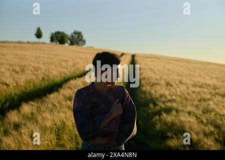 Una giovane donna si trova in un campo dorato al tramonto, guardando le spalle con un'espressione sorridente. Foto Stock