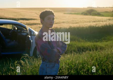 Una giovane donna si trova accanto a un'auto blu in un campo di erba alta dorata, con il sole che tramonta alle spalle e che emette un caldo bagliore. Foto Stock