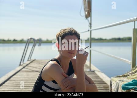 Una giovane donna è seduta su un molo di legno vicino a un lago, fumando una sigaretta, con una postura rilassata sotto un cielo azzurro. Foto Stock