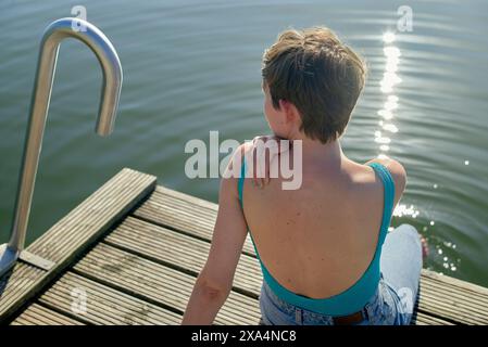 Una giovane donna siede su un molo di legno accanto a una scala di metallo, guardando l'acqua, con la luce del sole che si riflette sulla superficie. Foto Stock