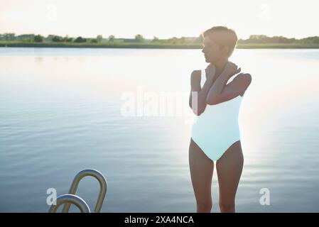 Una donna si trova accanto al lago al crepuscolo, indossando un costume da bagno bianco monopezzo, che sembra penosa mentre guarda sull'acqua con le braccia incrociate. Foto Stock