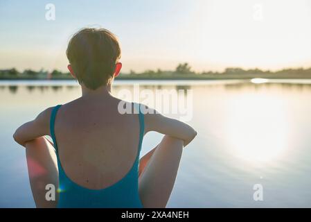 Una giovane donna è seduta accanto a un tranquillo lago al tramonto, con la schiena verso la macchina fotografica e le mani appoggiate sul retro della testa. Foto Stock