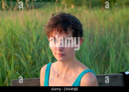Una giovane donna con i capelli scuri corti e una canotta blu si siede di fronte all'erba verde alta durante l'ora d'oro, guardando direttamente la fotocamera con un'espressione calma. Foto Stock