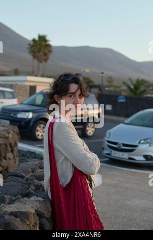Una giovane donna sorridente è in piedi all'aperto con auto parcheggiate e una montagna sullo sfondo durante quello che sembra essere il tardo pomeriggio. Foto Stock