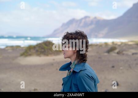 Una giovane donna con i capelli ricci sta in piedi su una spiaggia con montagne sullo sfondo, guardando in lontananza. Foto Stock