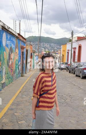 Una donna sorride mentre si trova su una strada acciottolata con edifici colorati e Street art sullo sfondo, con una collina in lontananza. Foto Stock