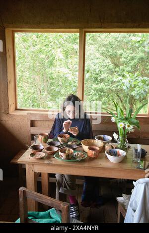 Una giovane donna è seduta ad un tavolo rustico in legno in una sala accogliente con vista sul verde esterno, gustando un pasto da una vasta gamma di ciotole in ceramica. Foto Stock