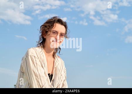 Una donna sorridente con i capelli ricci che indossa occhiali e una camicia a righe chiare si staglia su uno sfondo blu chiaro. Foto Stock
