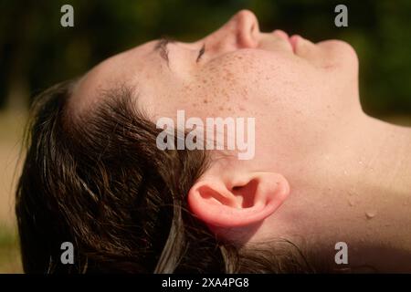 Primo piano del profilo di una persona con particolare attenzione all'orecchio e alla mandibola, su uno sfondo verde sfocato, con pelle baciata dal sole e gocce d'acqua. Foto Stock