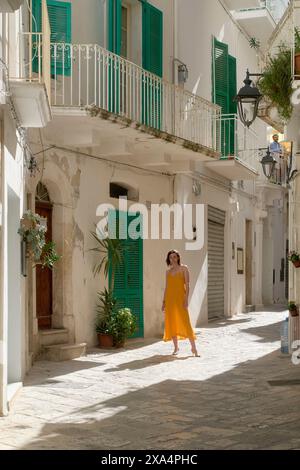 Una donna in un abito giallo che scorre passeggia lungo una pittoresca strada illuminata dal sole fiancheggiata da tradizionali edifici bianchi con persiane verdi. Foto Stock