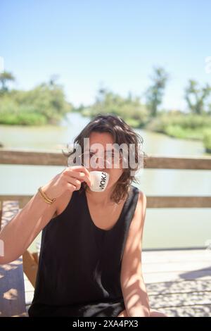 Una giovane donna seduta all'aperto a un tavolo di legno, bevendo da una tazza bianca con testo su di essa, con un fiume sereno e verde sullo sfondo. Foto Stock