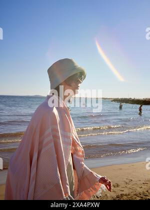 Una giovane donna sta sulla spiaggia drappeggiata da una coperta a righe rosa, indossa un cappello da secchio, con il sole che proietta una calda luce e un bagliore di lenti. Foto Stock