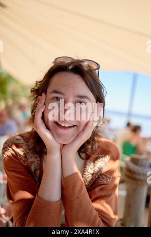 Una donna sorride alla macchina fotografica, poggia il mento sulle mani, con un ambiente all'aperto soleggiato sullo sfondo. Foto Stock