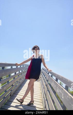 Una donna sta su un ponte di legno con la mano sulla ringhiera e una sciarpa rossa sulla spalla, sotto un cielo azzurro. Foto Stock