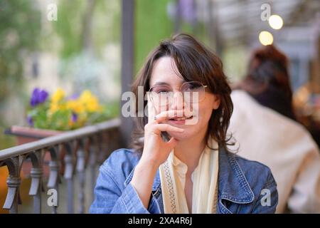 Una donna sorridente con gli occhiali è seduta in un bar all'aperto, appoggiando il mento sulla sua mano. Foto Stock