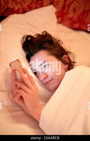 Giovane donna che si rilassa a letto mentre naviga sul suo smartphone Foto Stock