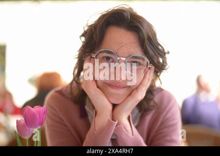 Donna sorridente con occhiali rotondi che appoggiano il mento sulle mani accanto a un tulipano rosa Foto Stock