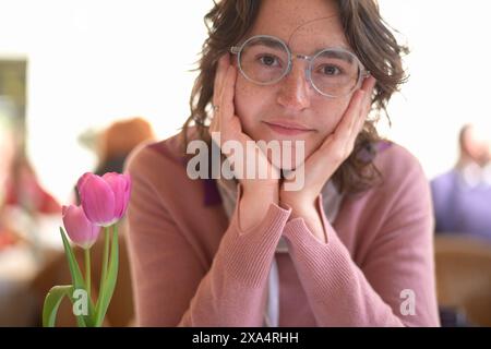 Donna sorridente con gli occhiali che appoggiano il mento sulle mani vicino a un tulipano rosa Foto Stock