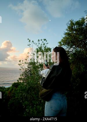 Una donna scatta una foto con il suo smartphone al tramonto in riva al mare circondato dal verde. Foto Stock