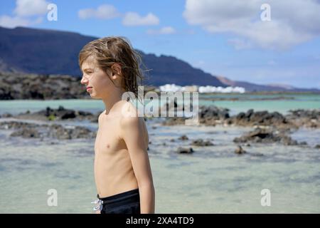 Il giovane ragazzo con i capelli spazzati dal vento si staglia davanti a un mare cristallino con formazioni rocciose e una costa serena sullo sfondo. Foto Stock