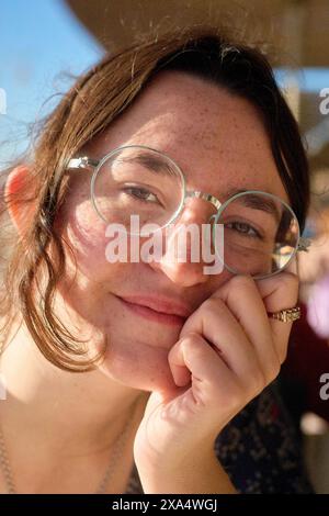 Giovane donna sorridente con gli occhiali che appoggiano il mento sulla mano in un ambiente illuminato dal sole. Foto Stock