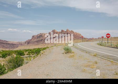 Strada curvilinea che conduce verso montagne rocciose desertiche sotto un cielo azzurro limpido con segnali di stop. Foto Stock