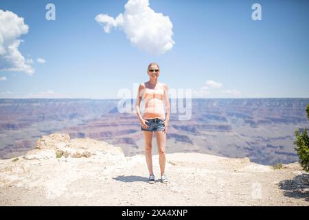 Donna sorridente in piedi ai margini del Grand Canyon in una giornata di sole con un cielo azzurro limpido e soffici nuvole in alto. Foto Stock