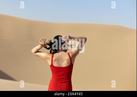 Donna con un vestito rosso in piedi davanti a una duna di sabbia con le mani che le tengono i capelli. Foto Stock