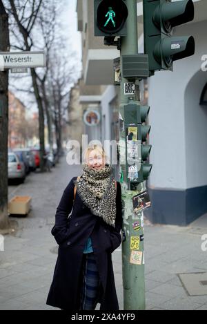 Donna sorridente in abbigliamento invernale in piedi accanto a un semaforo con un segnale pedonale verde in un ambiente urbano. Foto Stock