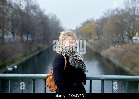 Donna con sciarpa e zaino in piedi su un ponte su un canale in una giornata nuvolosa. Foto Stock