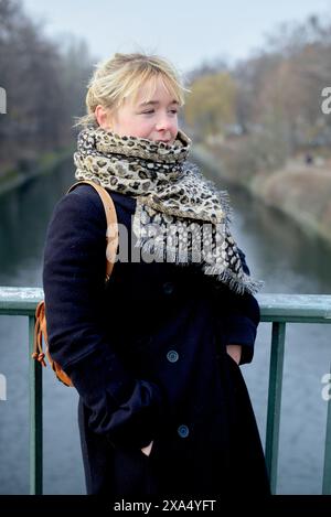 Una donna con un cappotto scuro e una sciarpa a motivi geometrici si erge su un ponte sul fiume, guardando di lato in una giornata nebbiosa. Foto Stock