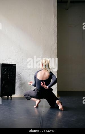 Una donna con una tuta grigia esegue una mossa di danza in uno spazioso studio con pareti bianche e un altoparlante nero sullo sfondo. Foto Stock