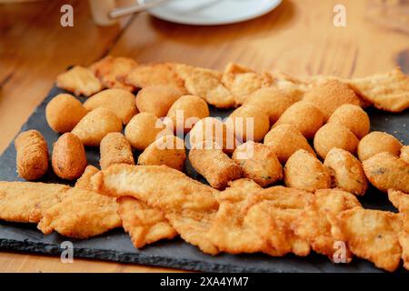 Un invitante piatto di spuntini fritti assortiti, tra cui crocchette dorate, strisce di pollo e polpette di formaggio, perfettamente disposte su un tabellone di ardesia Foto Stock