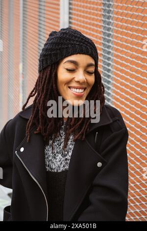 Giovane donna con dreadlock sorridente e appoggiata a una recinzione metalica per strada. Foto Stock