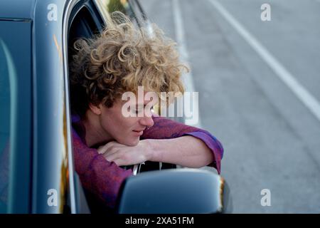 Uomo dai capelli ricci che si sporge dal finestrino dell'auto, godendosi la brezza. Foto Stock