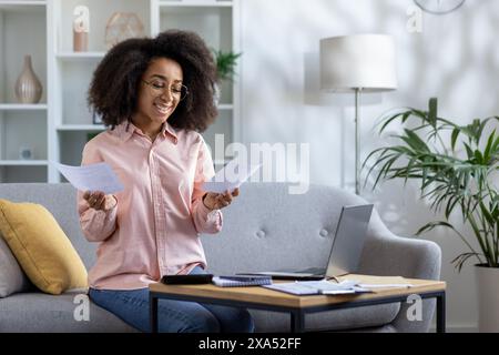 Donna sorridente che gestisce le finanze personali a casa, tiene in mano documenti e utilizza un computer portatile sul divano in salotto Foto Stock