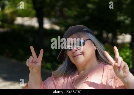 Una donna dai capelli grigi allegra e divertente, con cappello e occhiali da sole che fanno un segno di vittoria Foto Stock