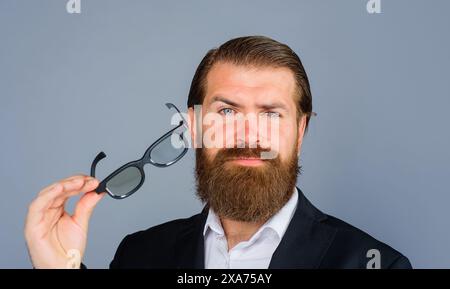 Il ritratto di un uomo barbuto serio si toglie gli occhiali da sole. Bell'uomo in tuta che si toglie gli occhiali da vista. Elegante uomo d'affari in abbigliamento formale con occhiali. Foto Stock