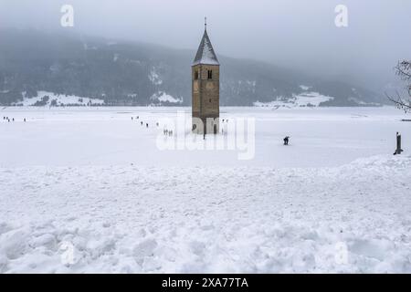 Un campanile in piedi nel mezzo del lago Resia durante l'inverno con persone che camminano intorno ad esso Foto Stock