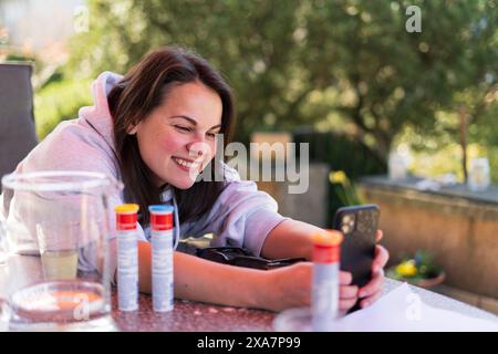 Una ragazza sorridente scatta una foto sul suo smartphone mentre legge sulla terrazza nel cortile di casa sua in estate. Concetto di felicità familiare Foto Stock