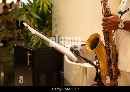 Un uomo suona un sassofono davanti a una pianta. Il sassofono è su un piedistallo accanto a uno spartito musicale Foto Stock