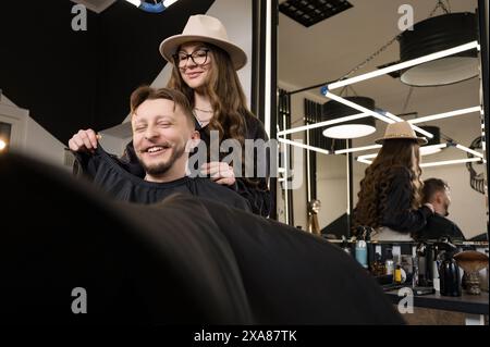 Un giovane con la barba in un mantello nero siede su una sedia in un barbiere. Il cliente sta aspettando un taglio di capelli e la rasatura della barba. Foto Stock