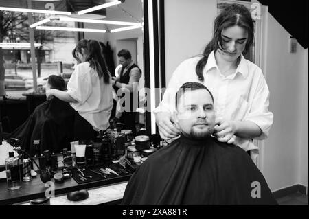 Un giovane con la barba in un mantello nero siede su una sedia in un barbiere. Il cliente sta aspettando un taglio di capelli e la rasatura della barba. Foto Stock