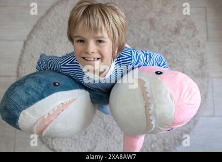 un bambino carino gioca con due grandi squali peluche che li tengono in due mani. Un grazioso ritratto di un bambino con i suoi giocattoli morbidi preferiti. Bambino comodo Foto Stock