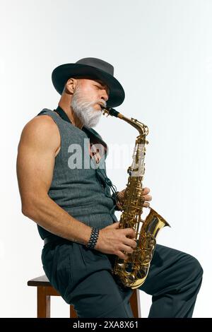 Uomo barbuto in gilet grigio senza maniche e cappello nero che suona jazz, sassofono dorato su sfondo bianco in studio. Foto Stock
