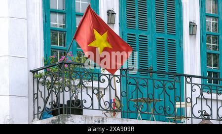 Una vibrante bandiera nazionale vietnamita rossa ondeggia in modo prominente su un balcone di una casa con porte e finestre blu in Hanoi City Street, simboleggiando la nazionale Foto Stock