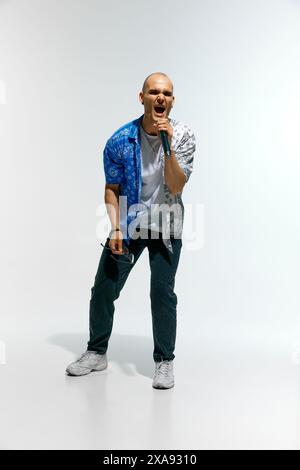 Abbigliamento casual da uomo completo di occhiali, si esibisce energicamente mentre canta in un microfono sullo sfondo bianco dello studio. Foto Stock