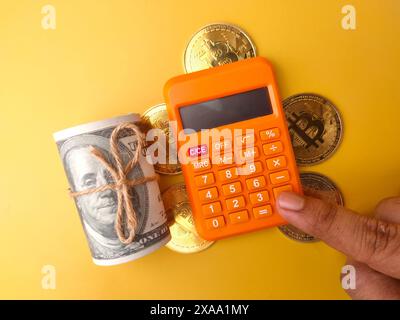 Qualcuno sta calcolando usando una calcolatrice con bitcoin dorato e banconote su sfondo giallo. Foto Stock