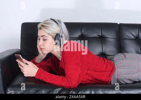 Giovane donna dai capelli biondi, con un maglione rosso e le cuffie, giace su un divano nero, guardando il suo telefono. Sembra rilassata e assorbita in h Foto Stock