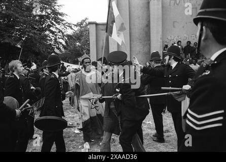 La rivolta del carnevale di Notting Hill agosto festivi lunedì 1976. Un gruppo di Rastafariani britannici neri che cantano e suonano tranquillamente. La polizia ha rotto il gruppo pensando che fossero dei rivoltosi. Alcuni poliziotti trasportano coperchi per la pattumiera da usare come scudo e hanno disegnato i loro manichini. Notting Hill, Londra, Inghilterra 30 agosto. 1970 UK HOMER SYKES Foto Stock